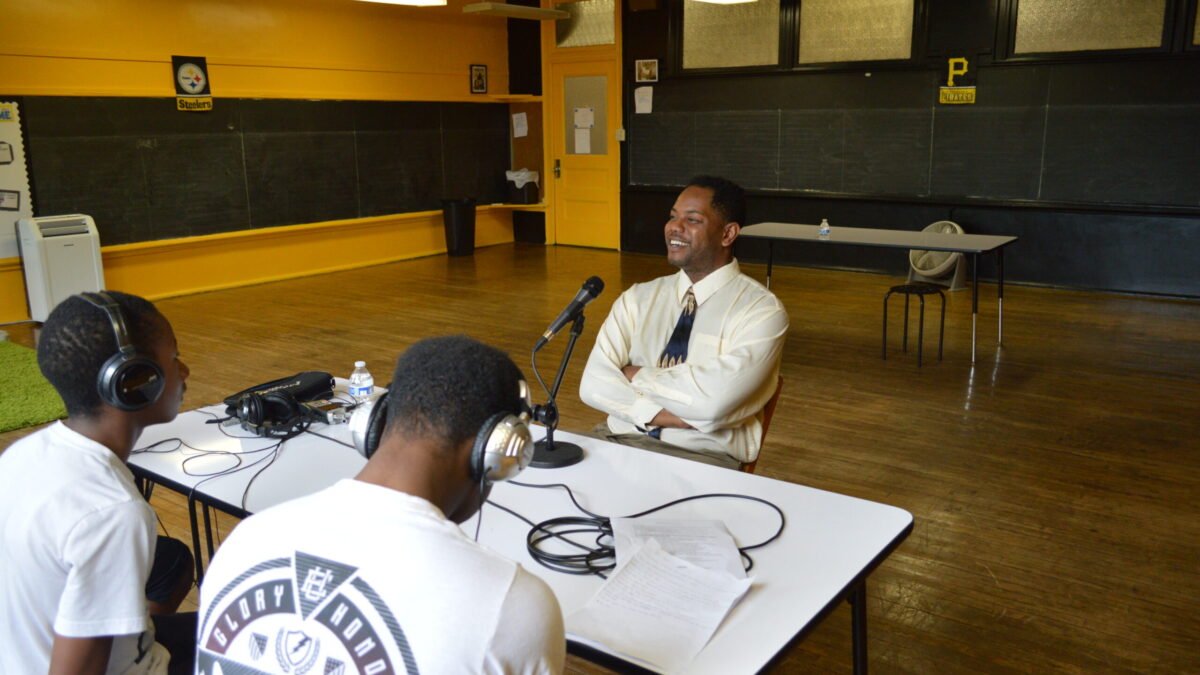 Two teens wearing headphones interviewing an adult speaking into a microphone connected to a portable recorder on a white table in a classroom