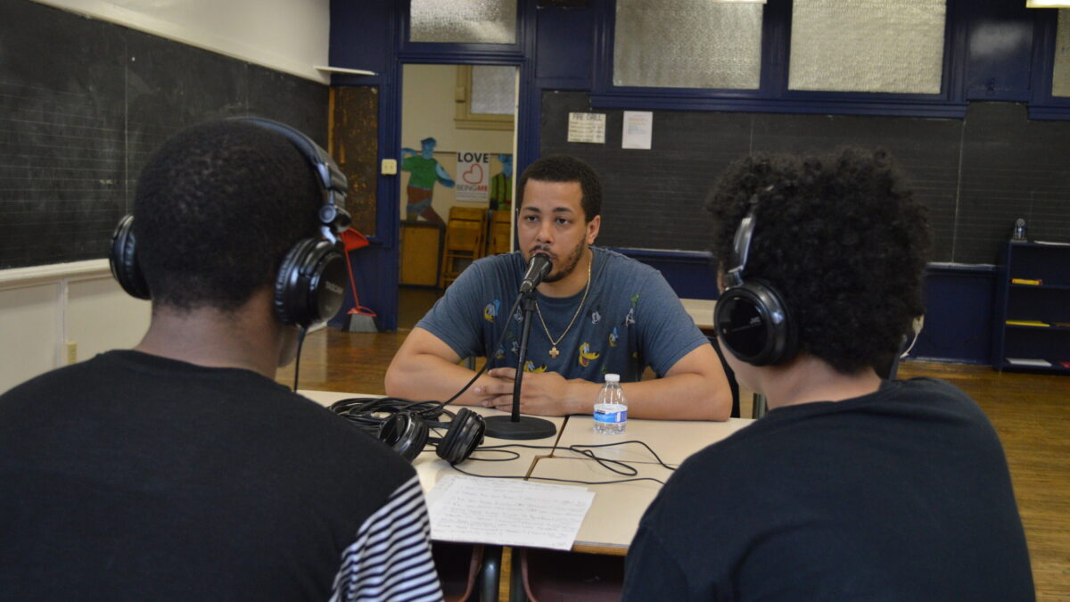 Two teens wearing headphones interviewing an adult speaking into a microphone connected to a portable recorder on two classroom desks in a classroom
