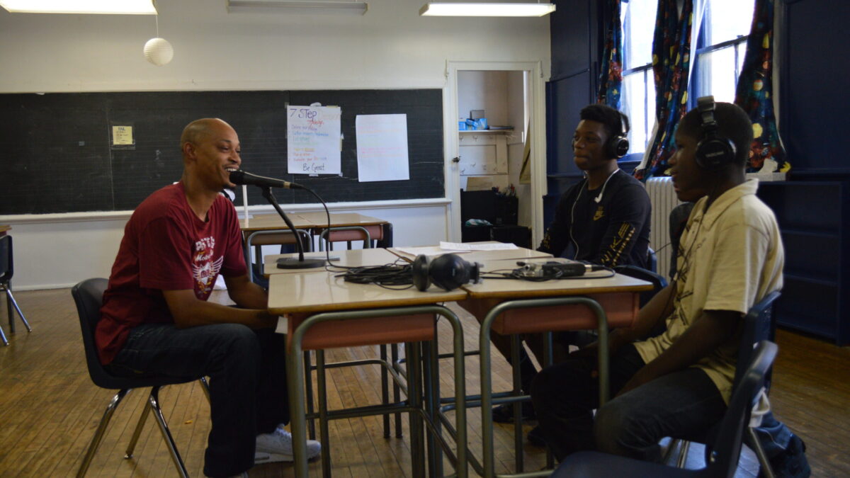 Three teens wearing headphones interviewing an adult speaking into a microphone connected to a portable recorder on two classroom desks in a classroom