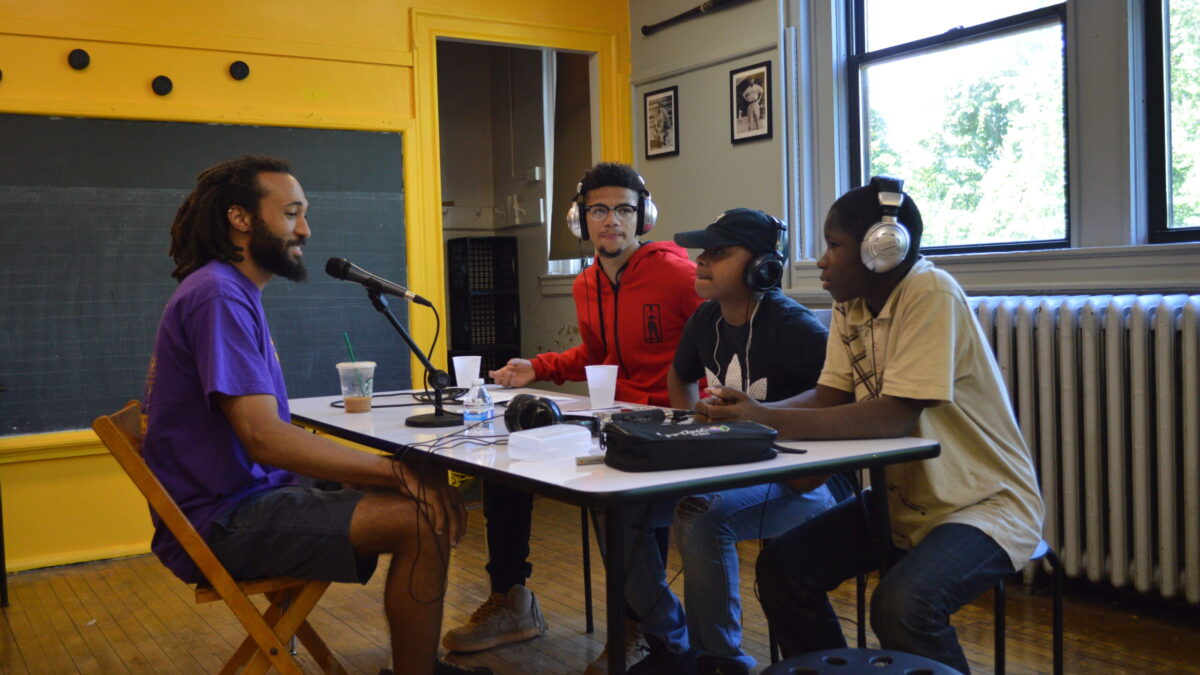 Three teens wearing headphones interviewing an adult speaking into a microphone connected to a portable recorder on a white table in a classroom