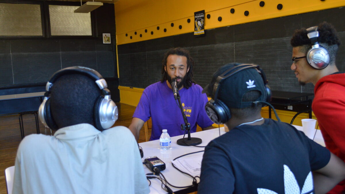 Three teens wearing headphones interviewing an adult speaking into a microphone connected to a portable recorder on a white table in a classroom