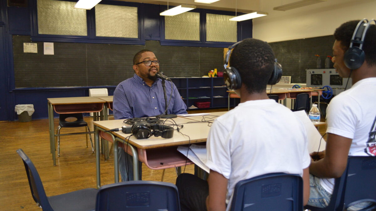 Two teens wearing headphones interviewing an adult speaking into a microphone connected to a portable recorder on two classroom desks in a classroom