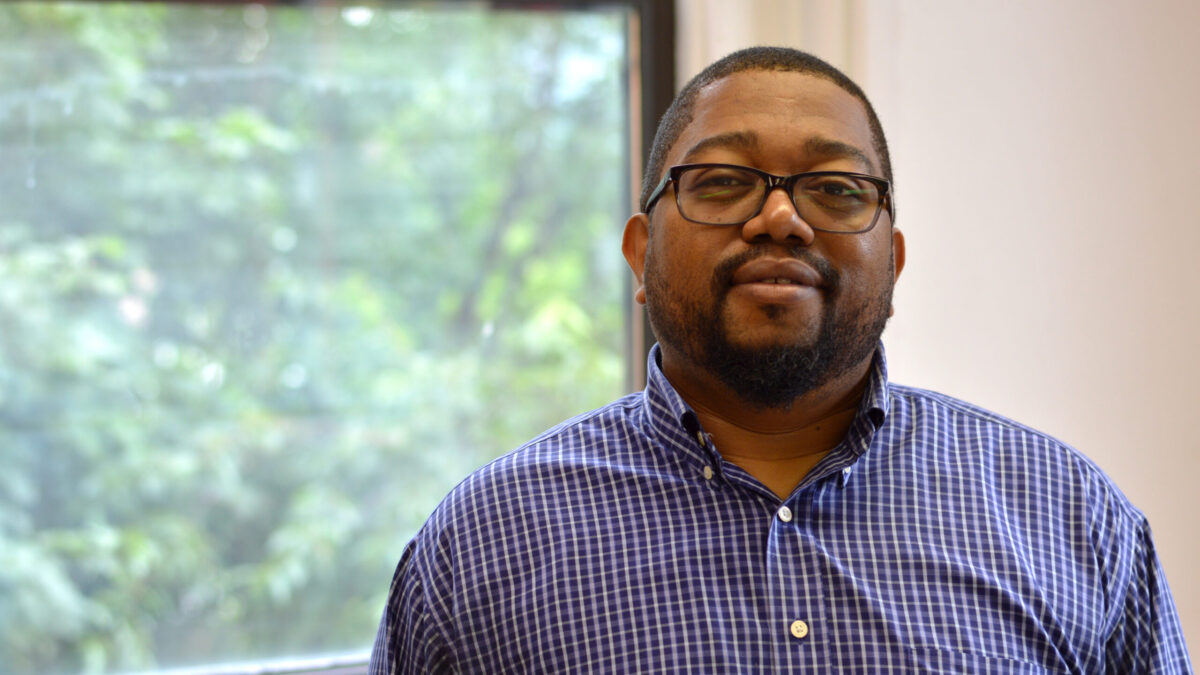 Adult with short black hair, a short black beard and mustache, glasses, and a blue and white checkered shirt posing in profile
