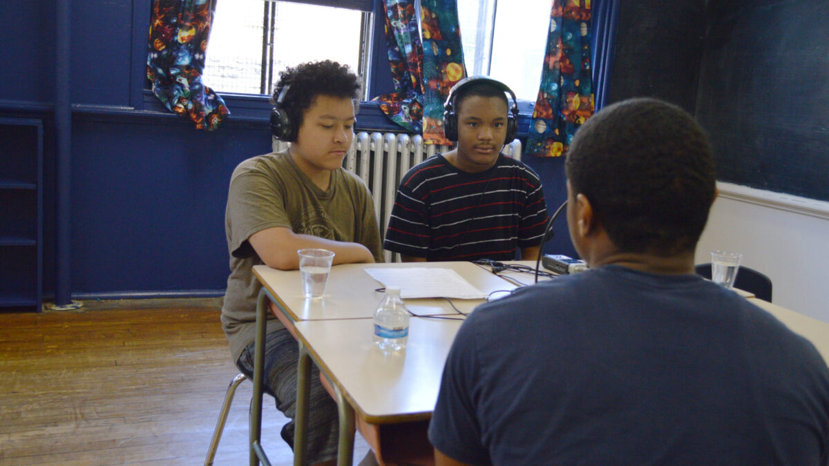 Two teens wearing headphones interviewing an adult speaking into a microphone connected to a portable recorder on two classroom desks in a classroom