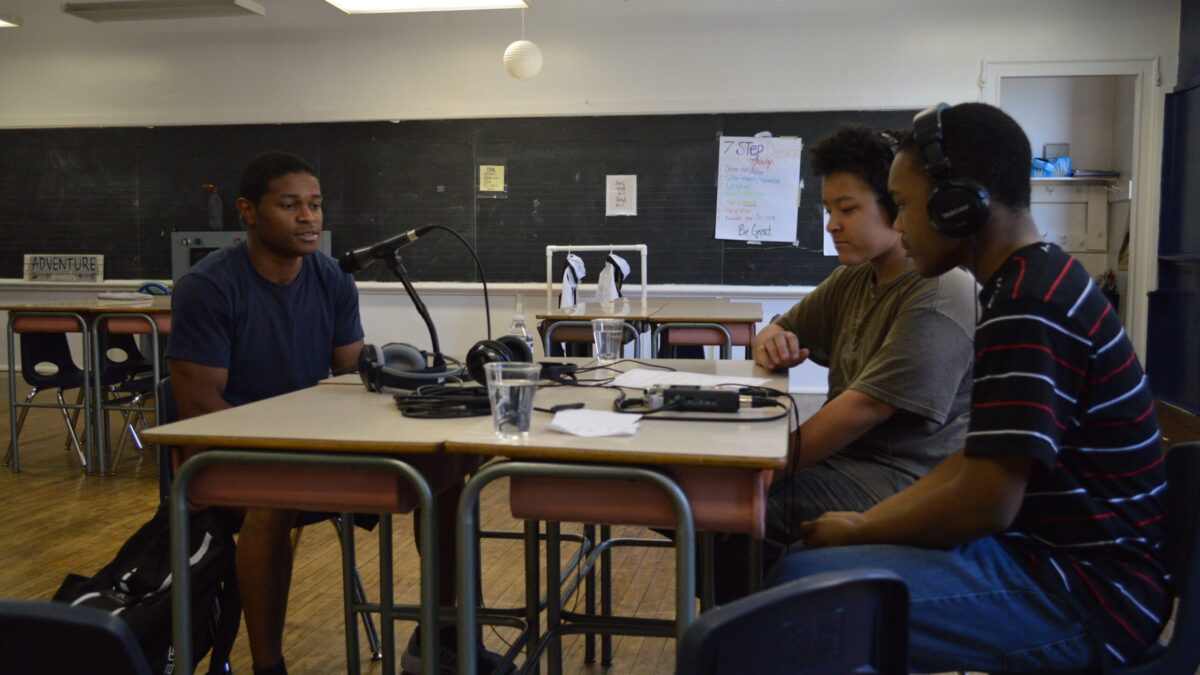 Two teens wearing headphones interviewing an individual speaking into a microphone connected to a portable recorder on two classroom desks in a classroom