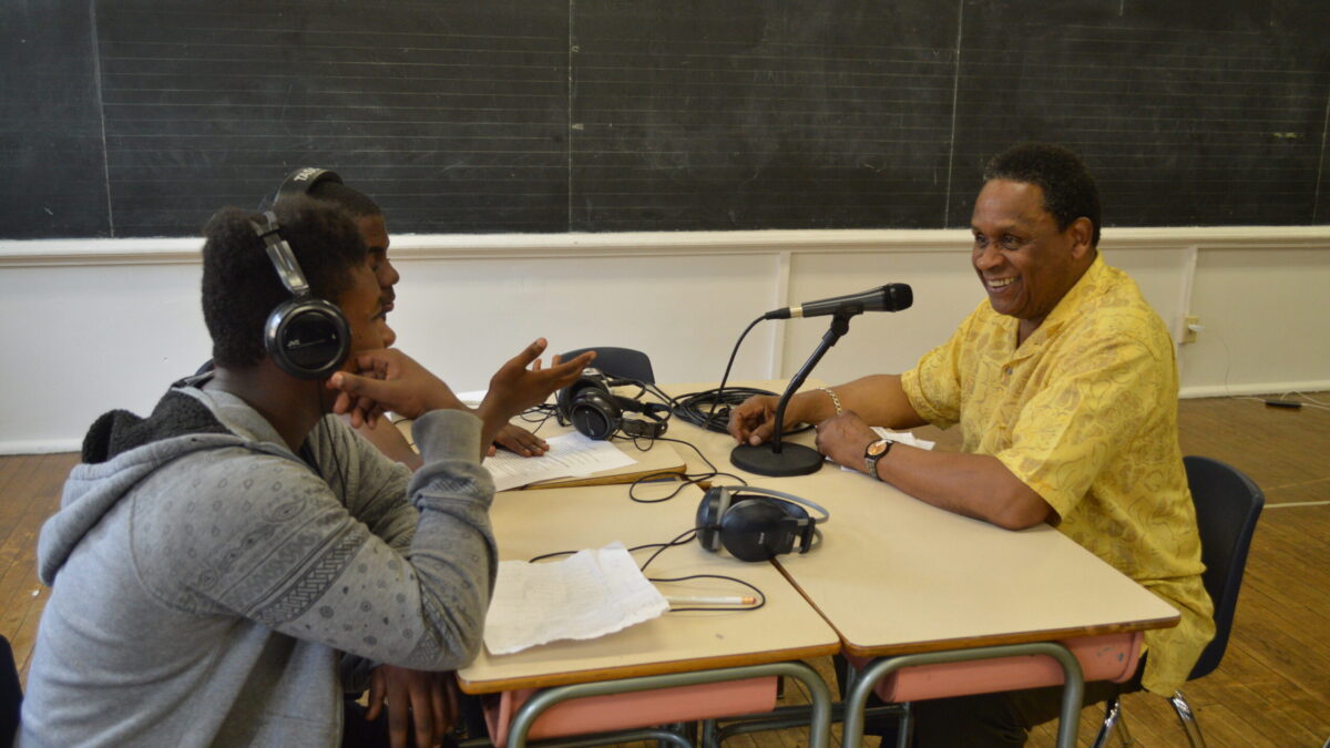 Two teens wearing headphones interviewing an adult speaking into a microphone connected to a portable recorder on two brown classroom desks in a classroom