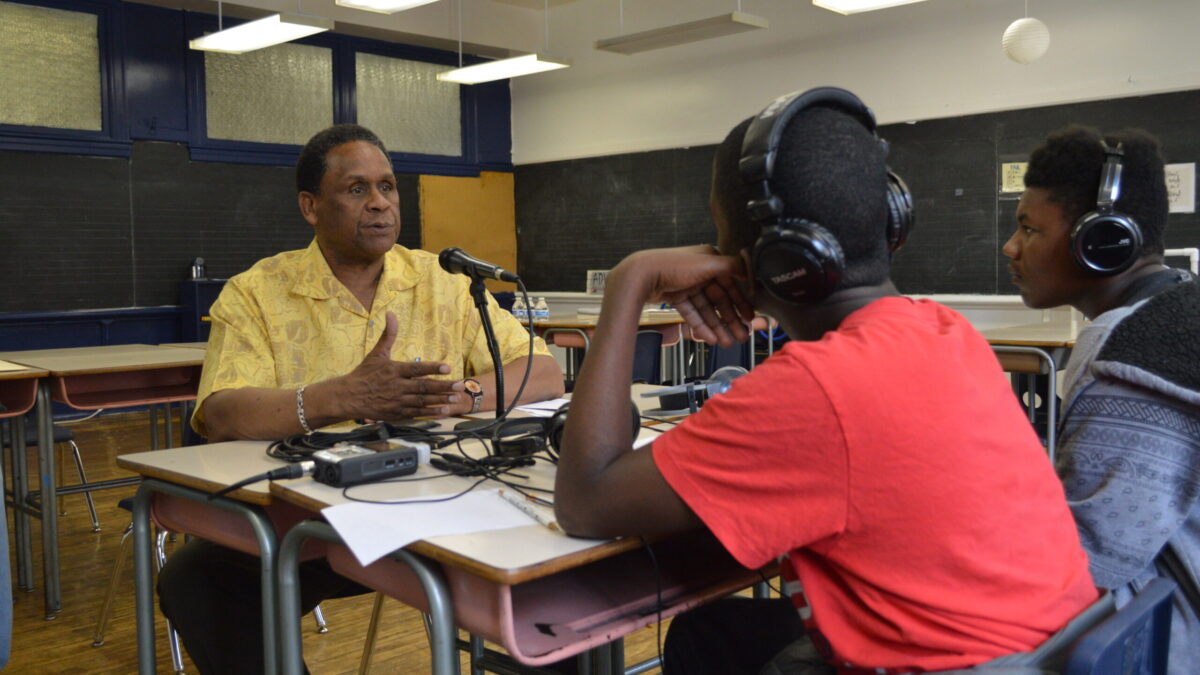 Two teens wearing headphones interviewing an adult speaking into a microphone connected to a portable recorder on two brown classroom desks in a classroom