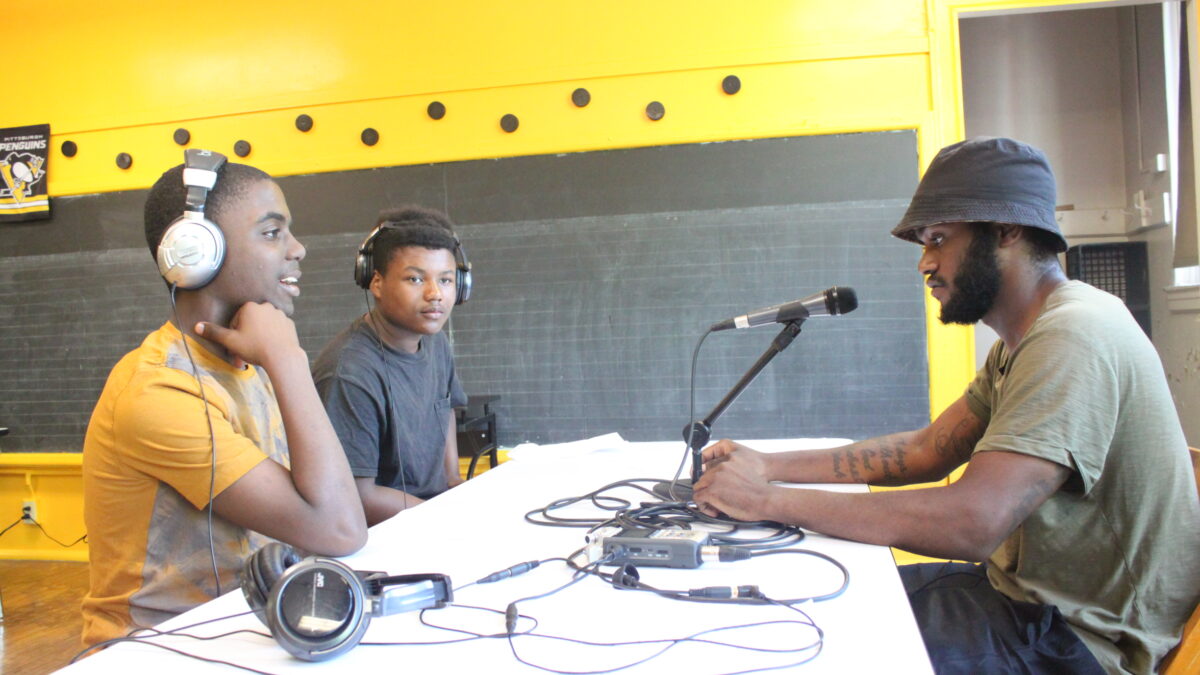 Two teens wearing headphones interviewing an adult speaking into a microphone connected to a portable recorder on a white table in a classroom