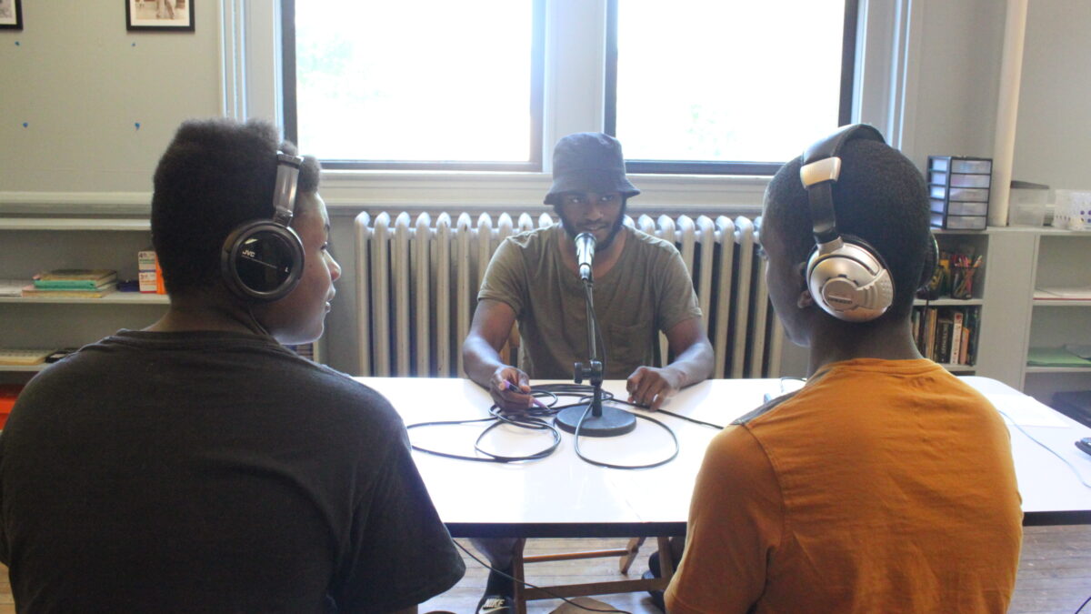 Two teens wearing headphones interviewing an adult speaking into a microphone on a white table in a classroom