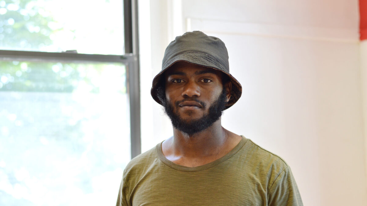 Adult with a short black mustache and beard wearing a black bucket hat and an olive green shirt posing in profile