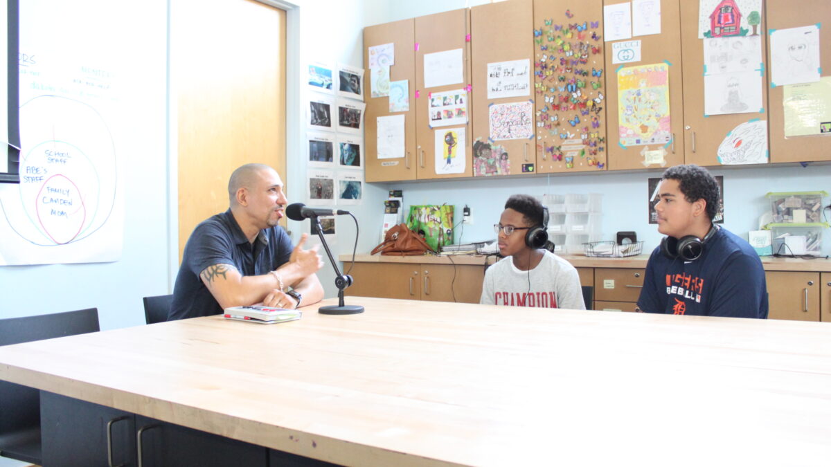 Two teens wearing headphones interviewing an adult speaking into a microphone on a table in a classroom