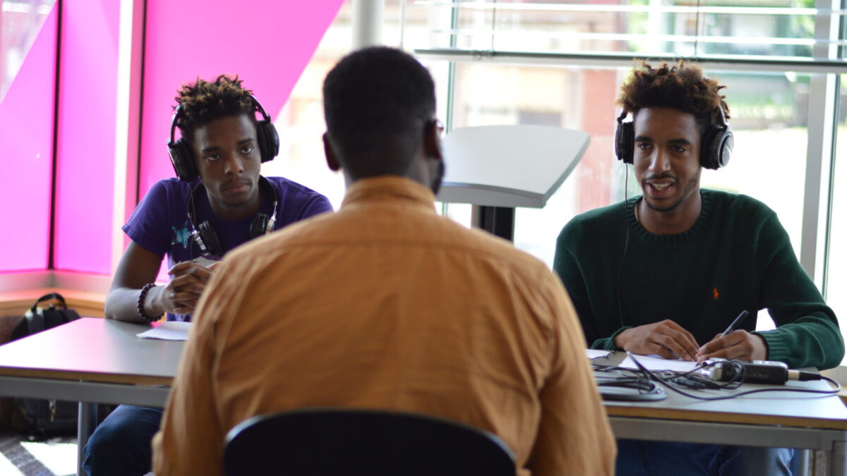 Two teens wearing headphones, one writing on a piece of paper, interviewing an adult sitting in front of a portable recorder on a brown table