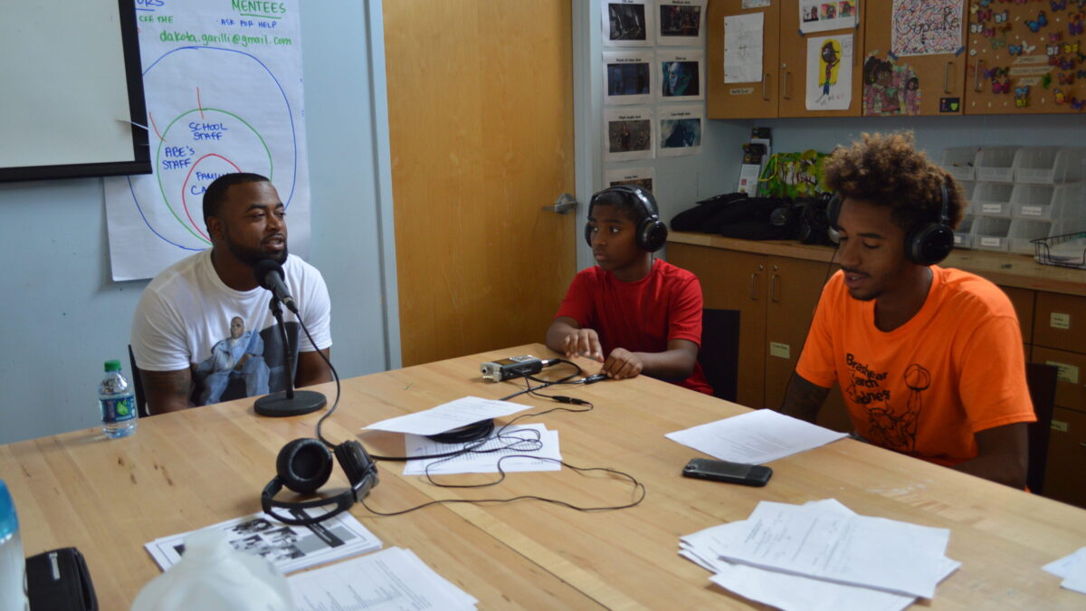 Two teens wearing headphones interviewing an adult speaking into a microphone connected to a portable recorder on a brown table