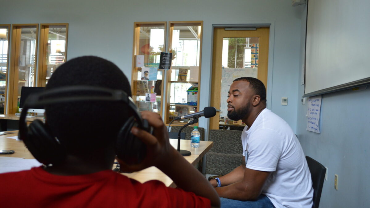 Teen wearing headphones interviewing an adult speaking into a microphone on a brown table