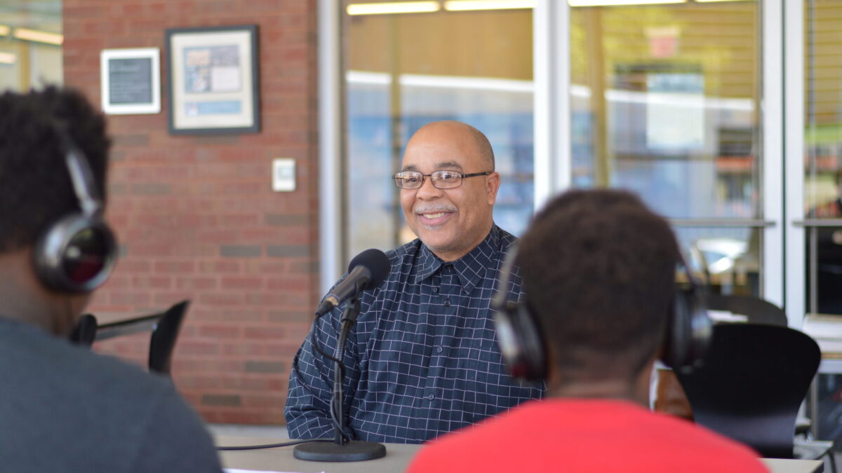 Two teens wearing headphones interviewing an adult speaking into a microphone on a brown table