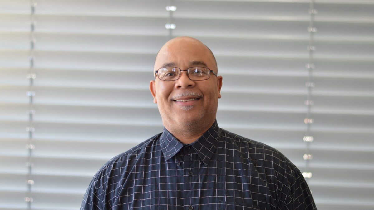 Bald adult with gray stubble and mustache and a blue and white checkered shirt smiling in profile