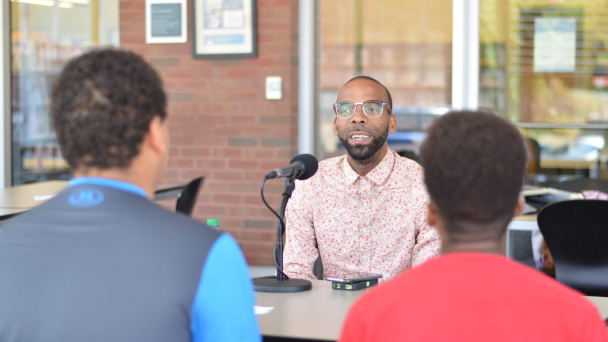 Two teens interviewing an adult speaking into a microphone on a brown table