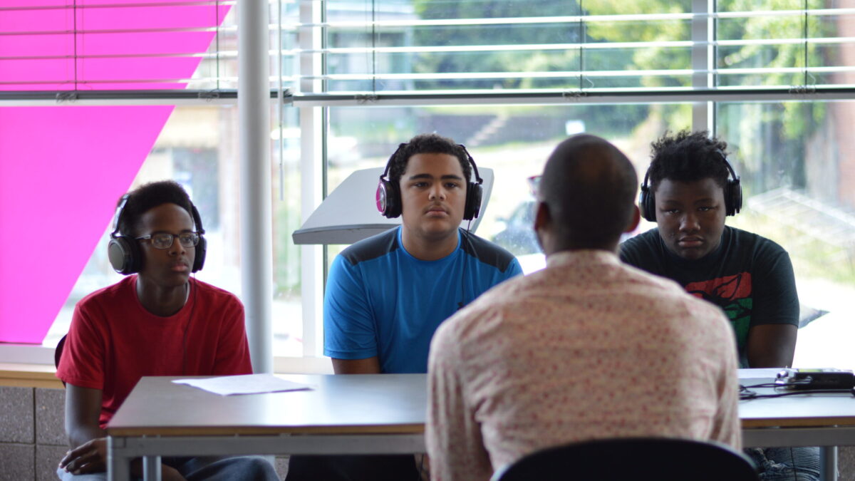 Three teens wearing headphones interviewing an adult sitting in front of a portable recorder on a brown table