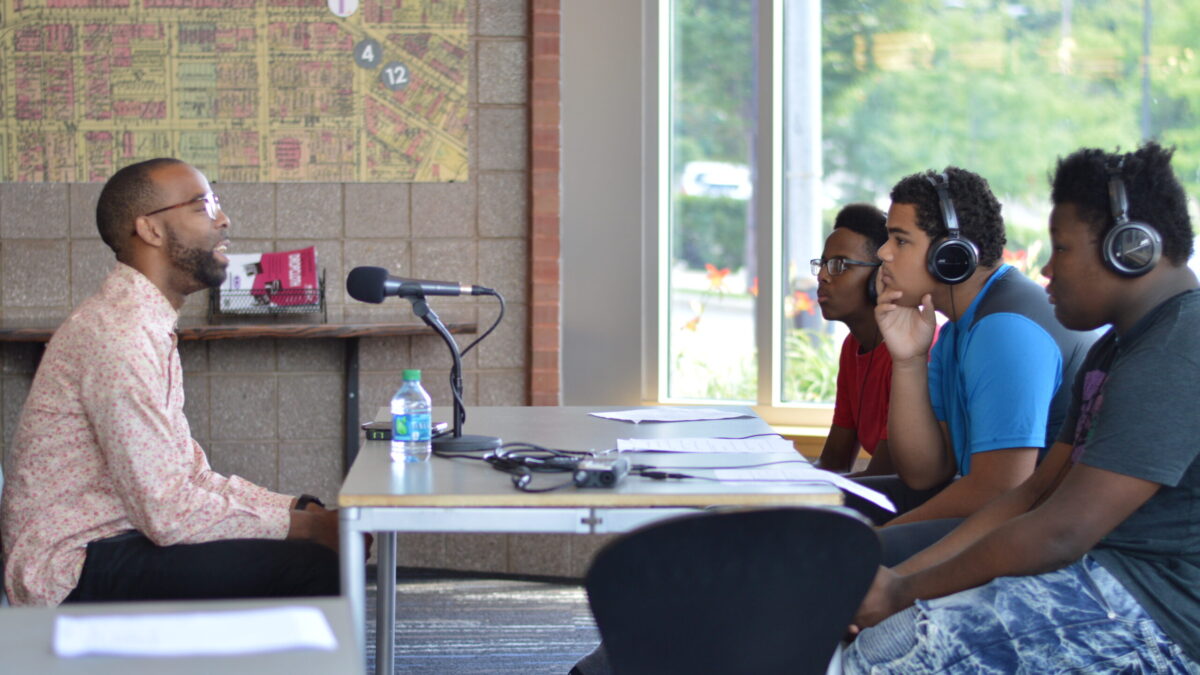 Three teens wearing headphones interviewing an adult speaking into a microphone connected to a portable recorder on a brown table