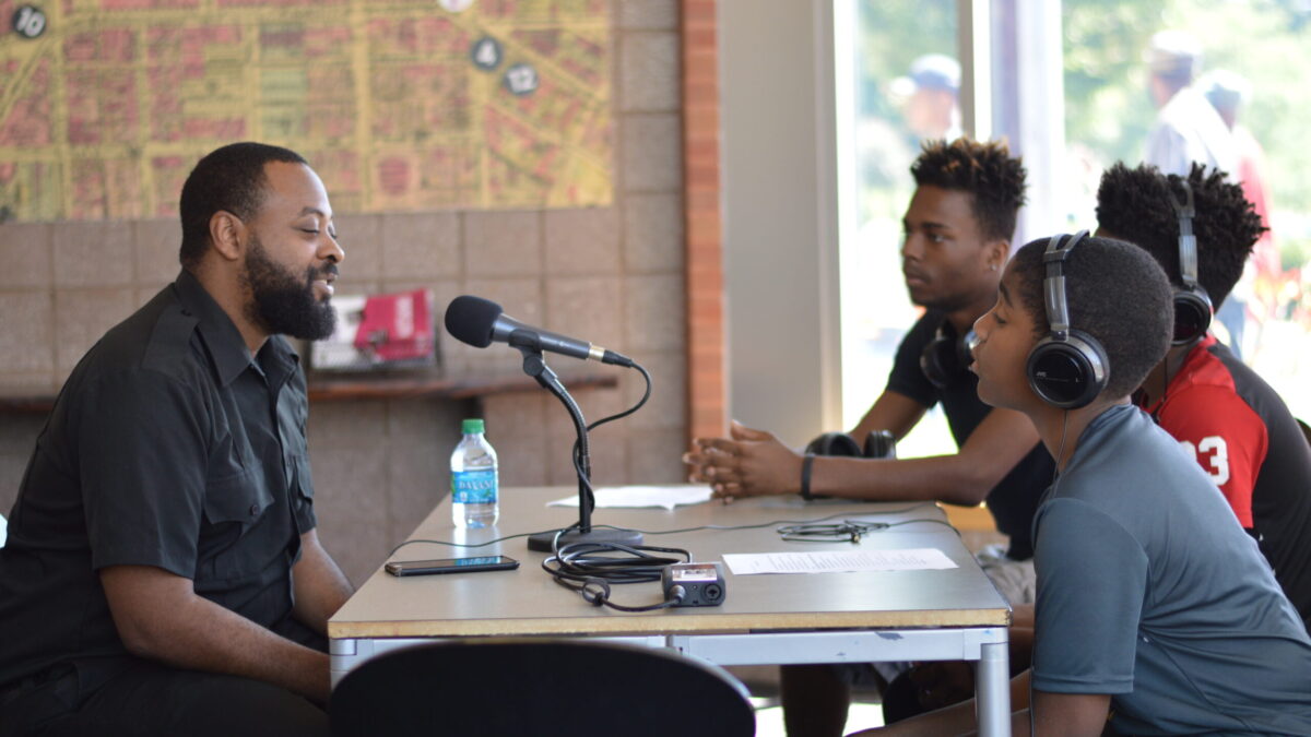 Three youth wearing headphones interviewing an adult speaking into a microphone connected to a portable recorder on a brown table