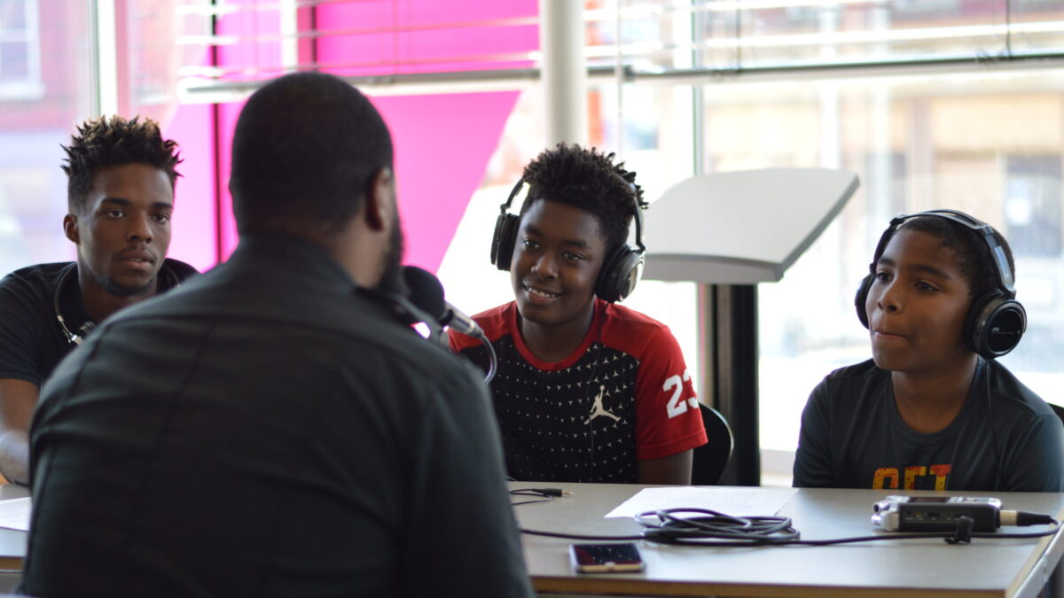 Three youth wearing headphones interviewing an adult speaking into a microphone connected to a portable recorder on a brown table