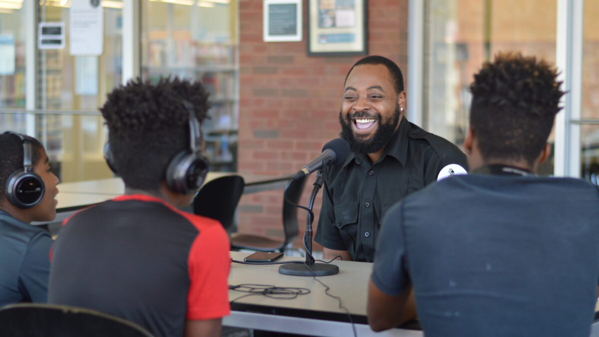 Three youth wearing headphones interviewing an adult speaking into a microphone on a table