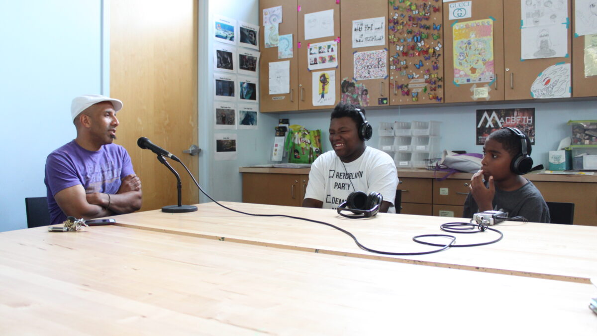Two teens wearing headphones interviewing an adult speaking into a microphone connected to a portable recorder on a brown table