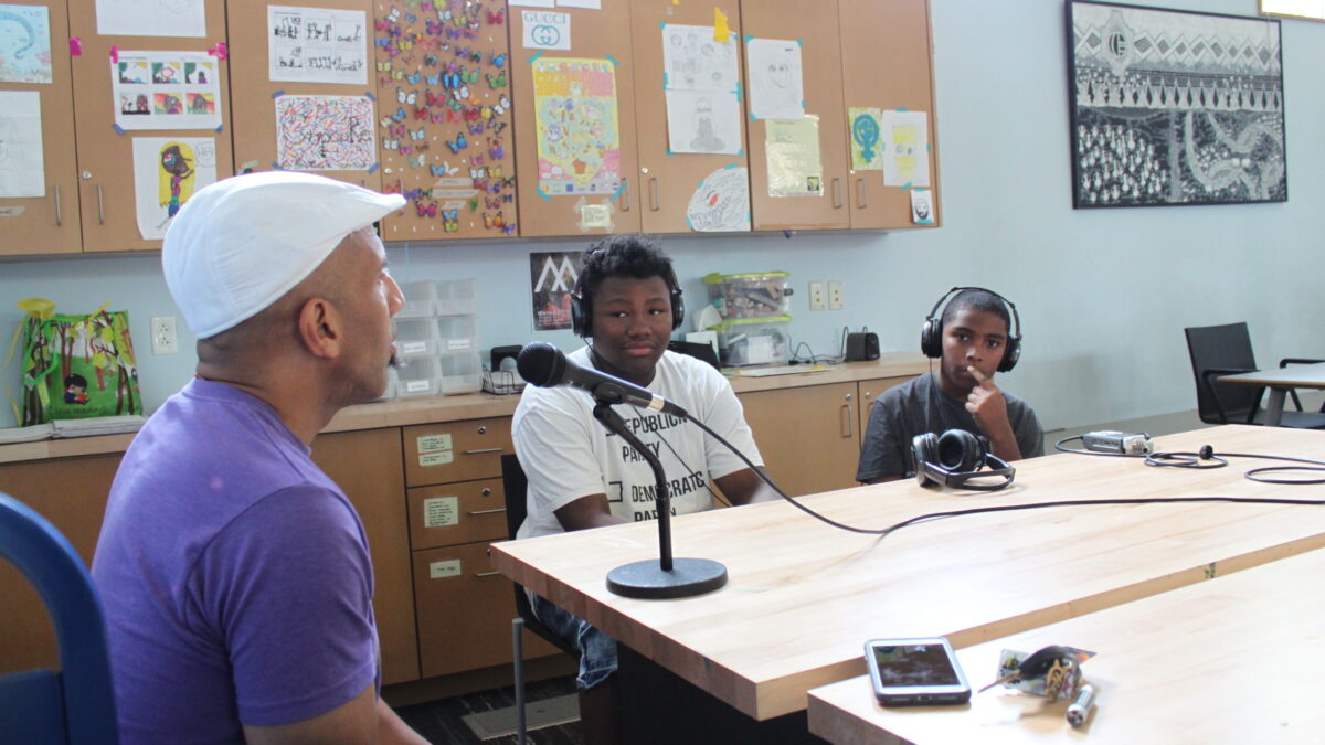 Two teens wearing headphones interviewing an adult speaking into a microphone connected to a portable recorder on a brown table