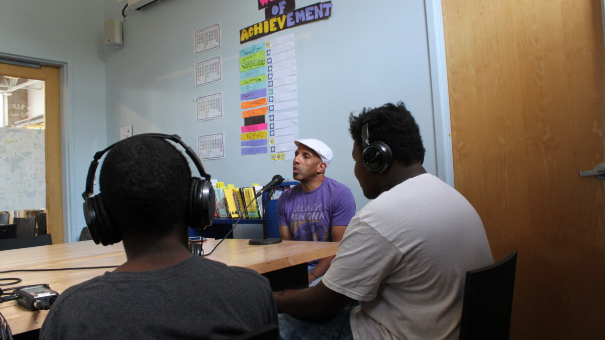 Two teens wearing headphones interviewing an adult speaking into a microphone connected to a portable recorder on a brown table