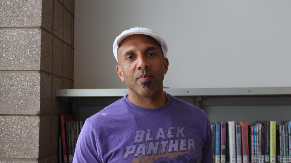 Adult with a white newsboy cap and a short black soul patch beard posing in profile
