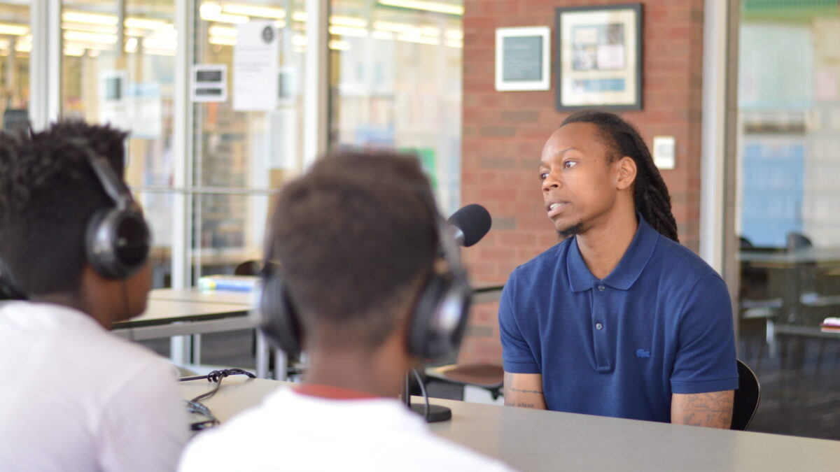 Two teens wearing headphones interviewing an adult speaking into a microphone on a brown table