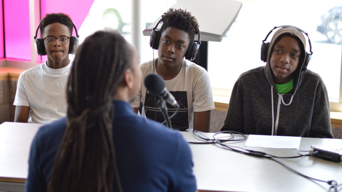 Three teens wearing headphones interviewing an adult speaking into a microphone connected to a portable recorder on a white table