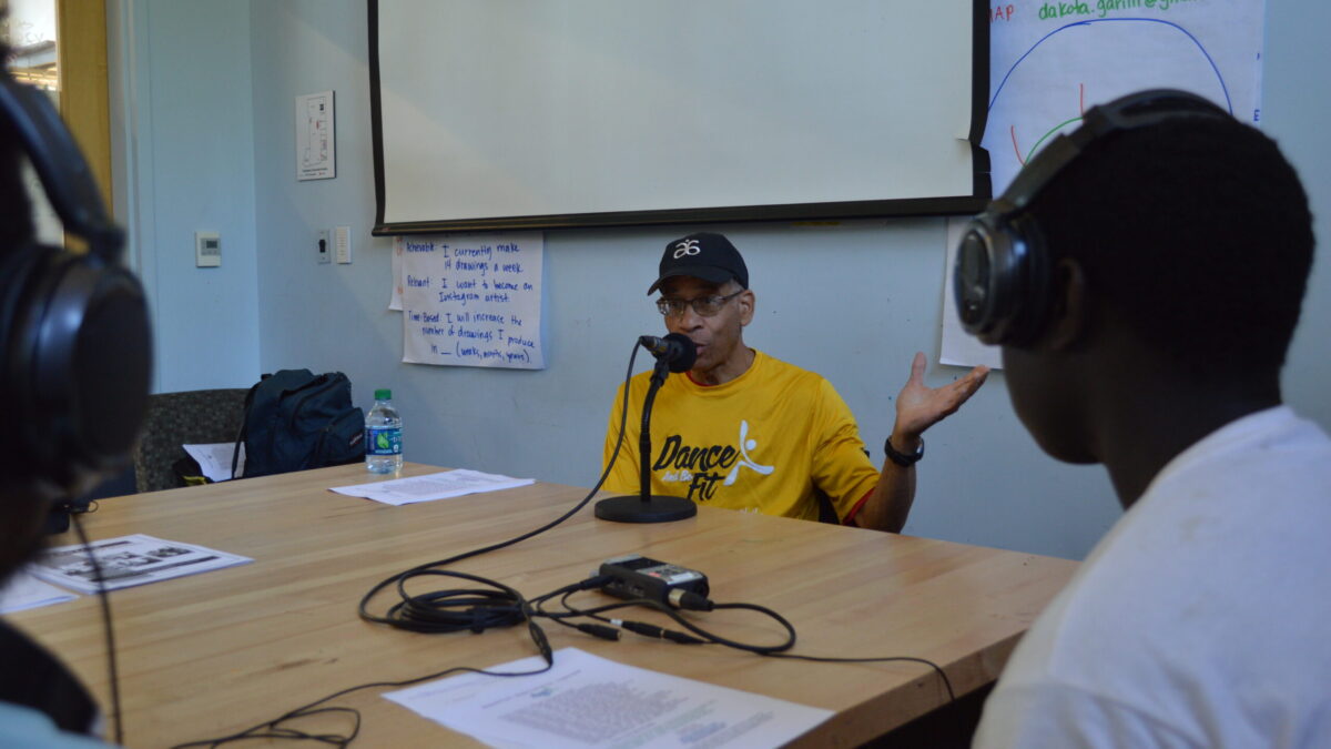 Two teens wearing headphones interviewing an adult speaking into a microphone connected to a portable recorder on a brown table