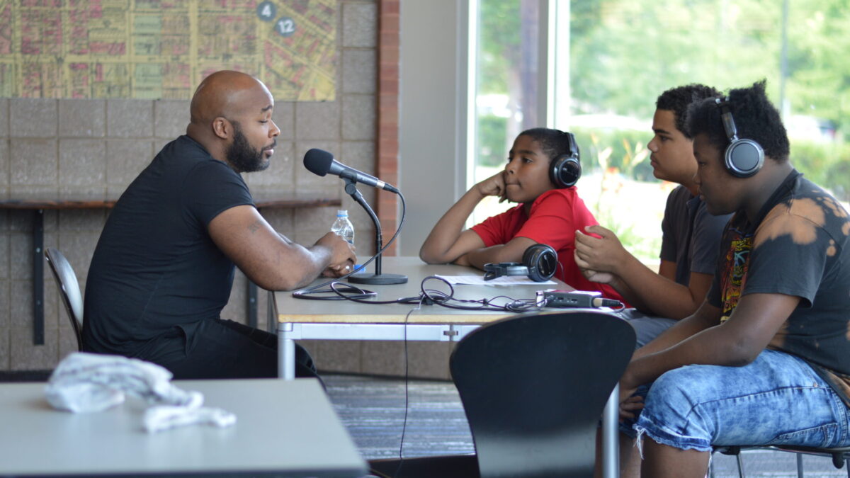 Three youth, two wearing headphones, interviewing an adult speaking into a microphone connected to a portable recorder on a brown table