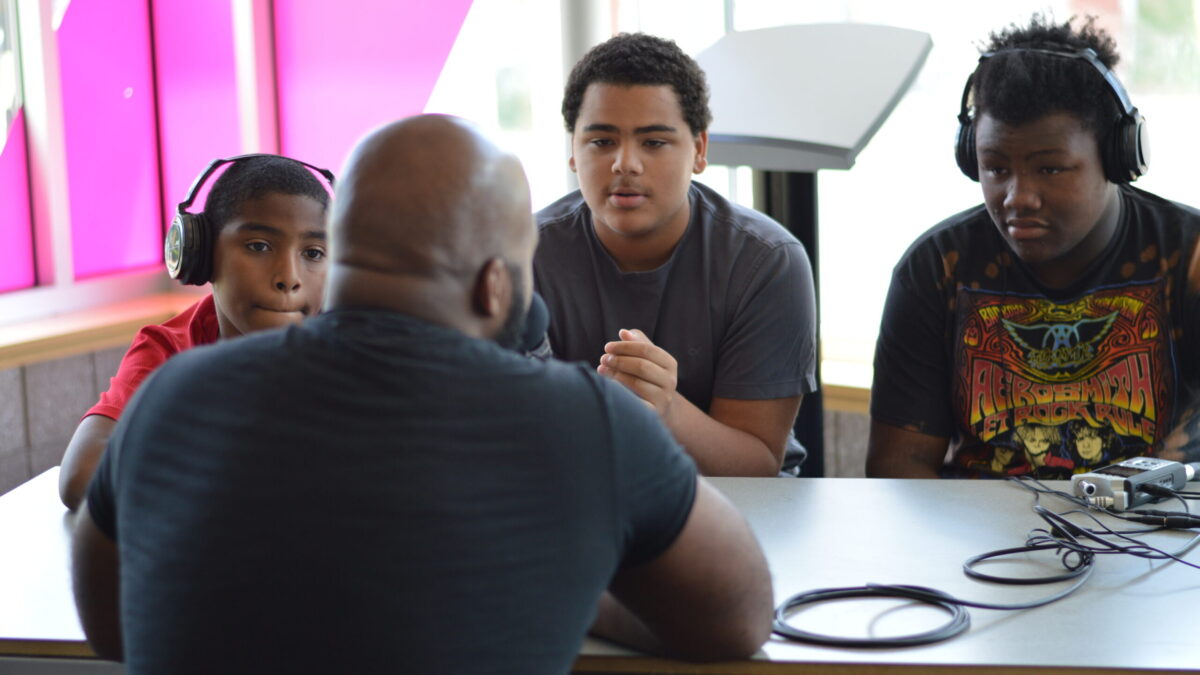 Three youth, two wearing headphones, interviewing an adult speaking into a microphone connected to a portable recorder on a brown table