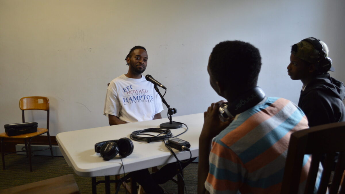 Two teens wearing headphones interviewing an adult sitting in front of a microphone connected to a portable recorder on a white table