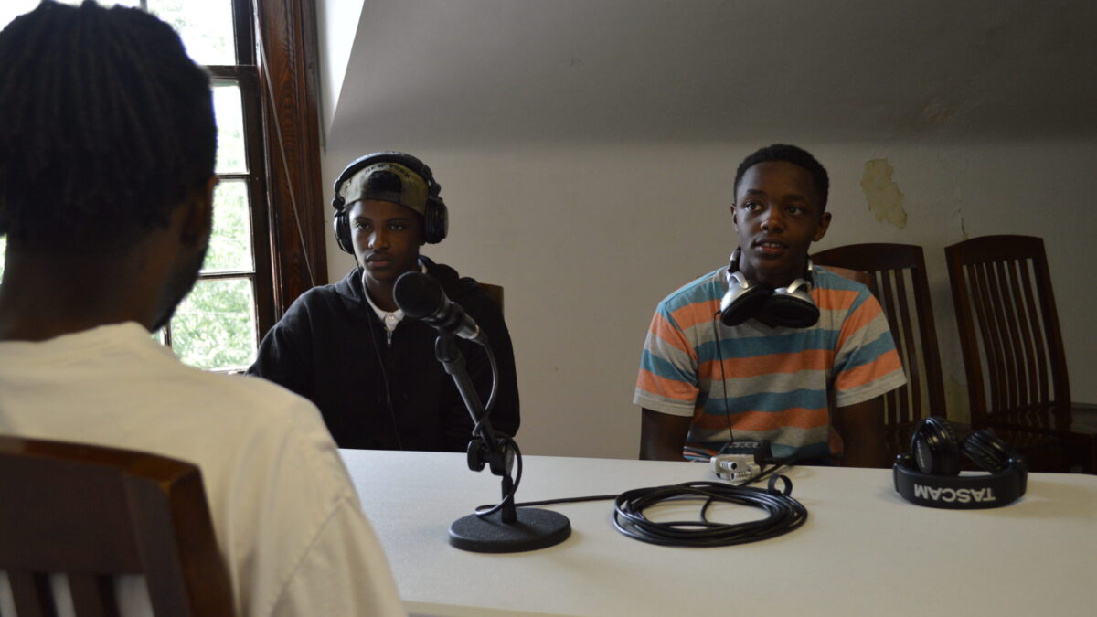 Two teens wearing headphones interviewing an adult sitting in front of a microphone connected to a portable recorder on a white table
