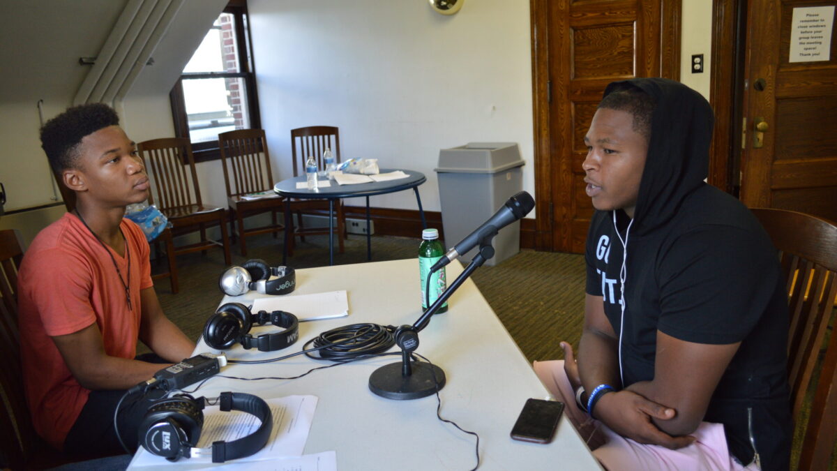 Teen interviewing an adult speaking into a microphone connected to a portable recorder on a white table