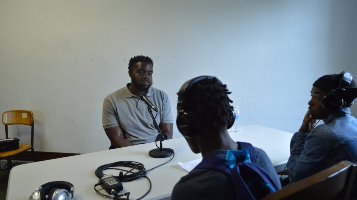 Two teens wearing headphones interviewing an adult sitting in front of a microphone connected to a portable recorder on a white table