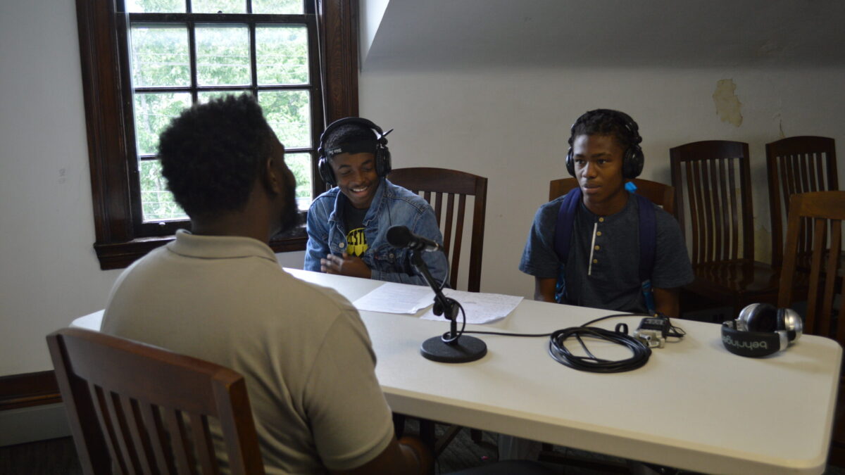 Two teens wearing headphones interviewing an adult speaking into a microphone connected to a portable recorder on a white table