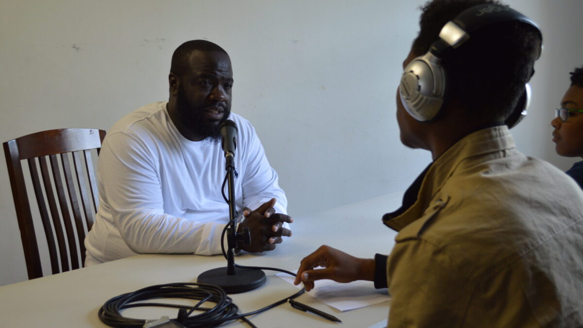 Two teens wearing headphones interviewing an adult speaking into a microphone connected to a portable recorder on a white table