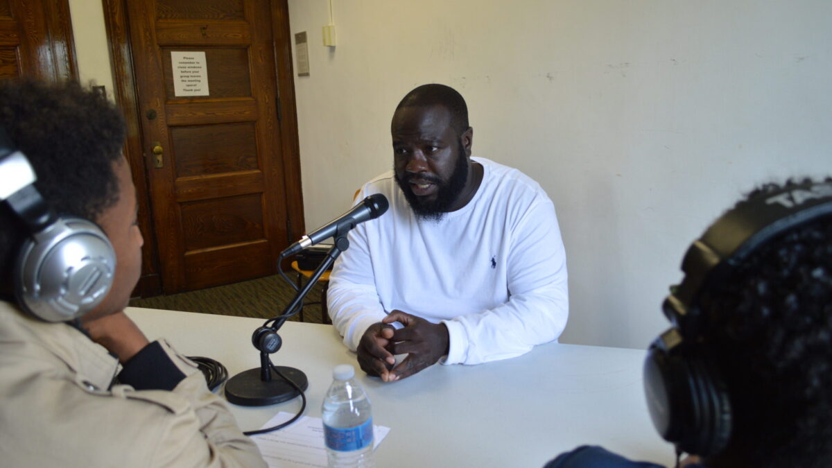 Two youth wearing headphones interviewing an adult speaking into a microphone on a white table