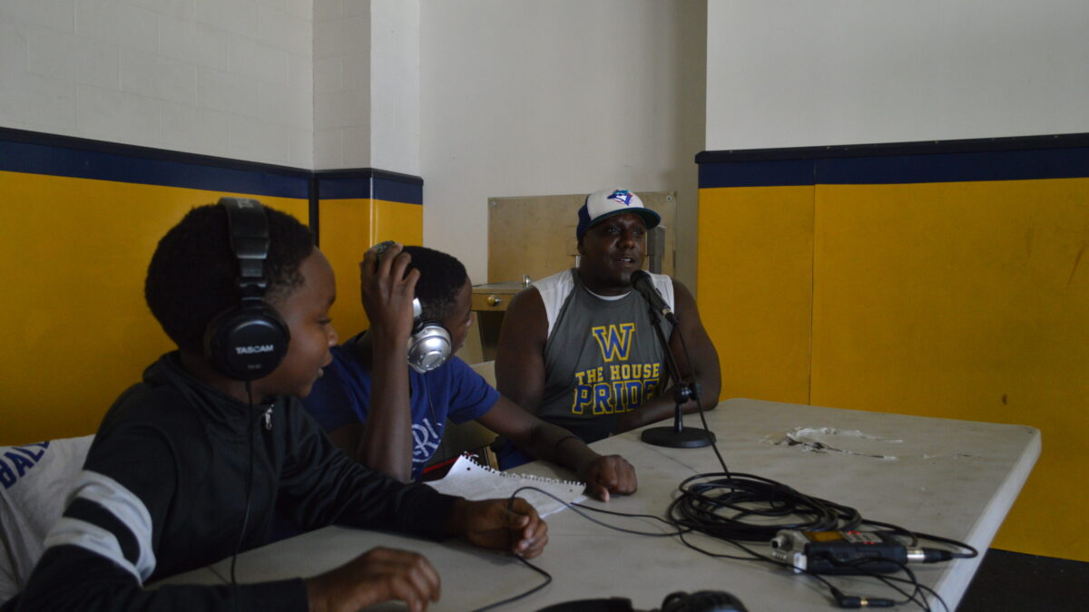Two youth wearing headphones interviewing an adult speaking into a microphone connected to a portable recorder on a white table