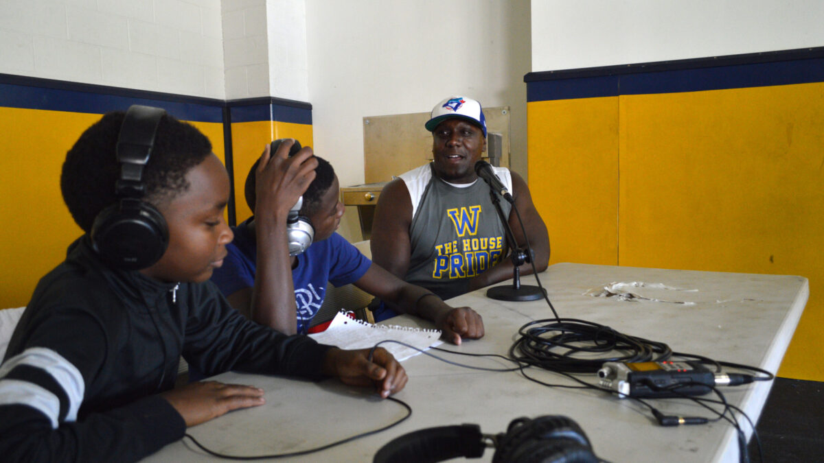Two youth wearing headphones interviewing an adult speaking into a microphone connected to a portable recorder on a white table