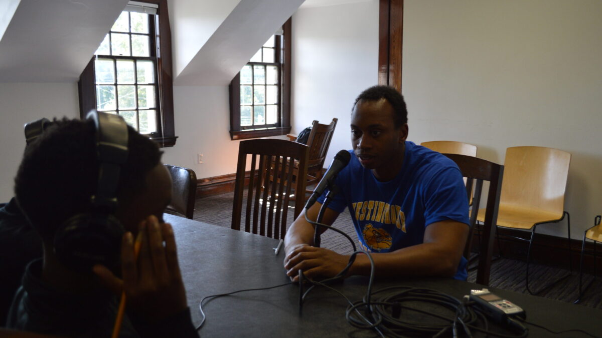 A youth wearing headphones interviewing an adult speaking into a microphone connected to a portable recorder on a black table