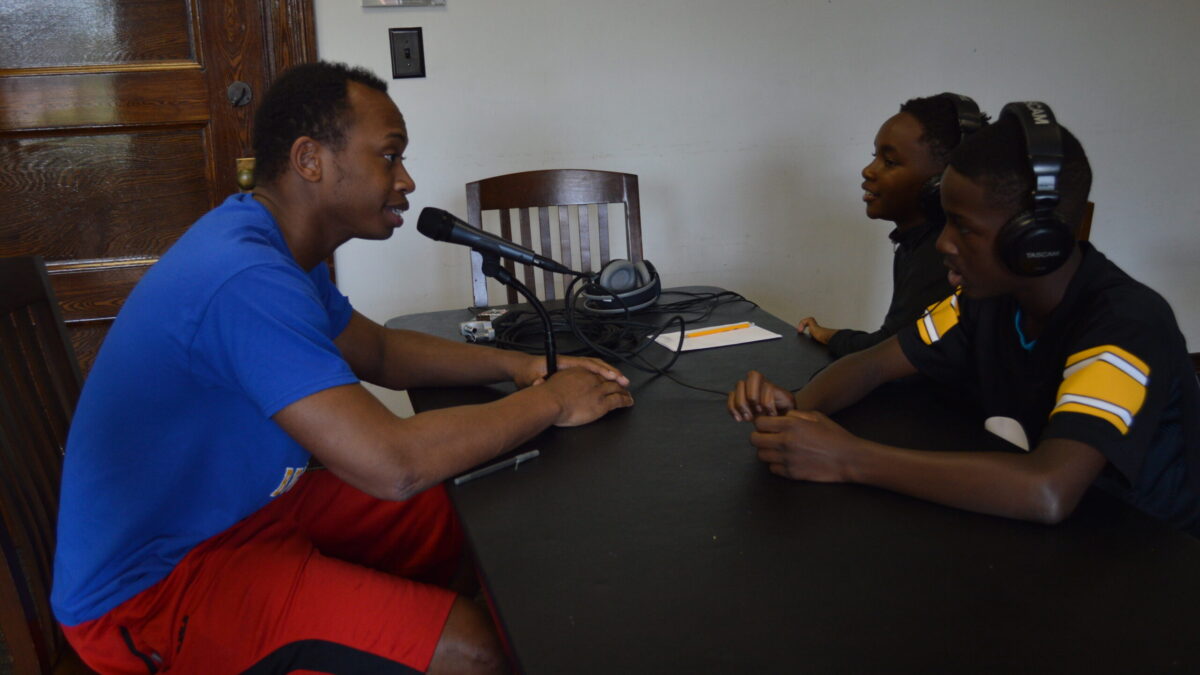 Two youth wearing headphones interviewing an adult speaking into a microphone connected to a portable recorder on a black table