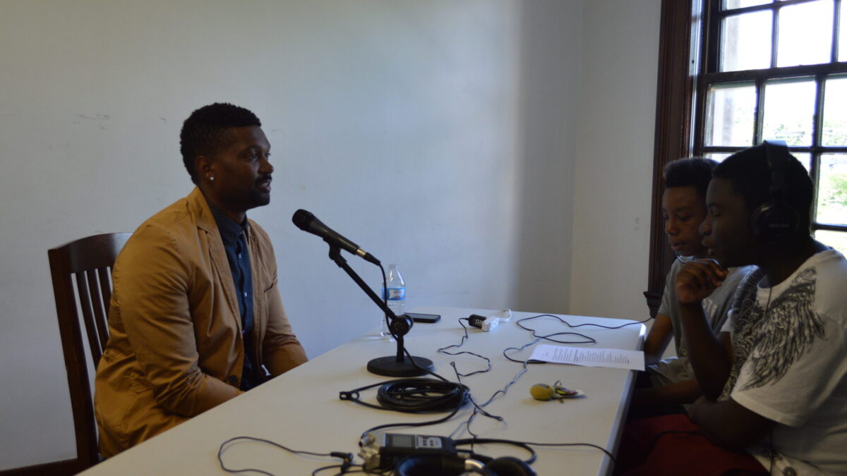 Two teens wearing headphones interviewing an adult speaking into a microphone connected to a portable recorder on a white table