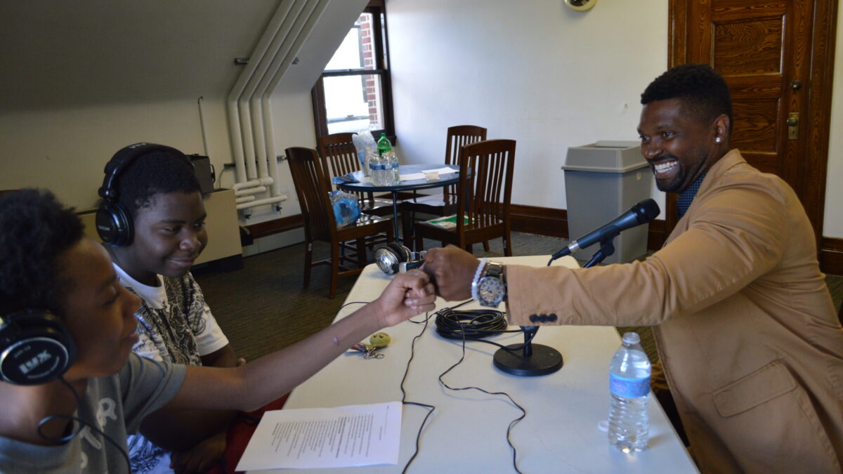 Two teens wearing headphones interviewing an adult speaking into a microphone connected to a portable recorder on a white table, the adult fist bumping one of the teens
