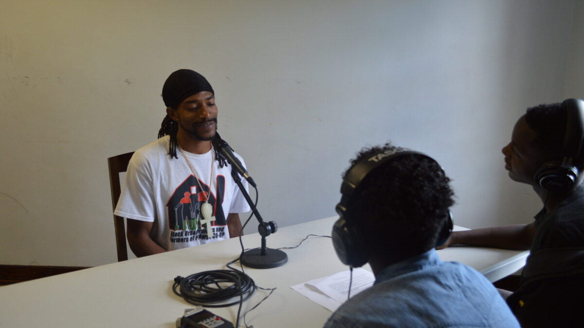 Two teens wearing headphones interviewing an adult speaking into a microphone connected to a portable recorder on a white table