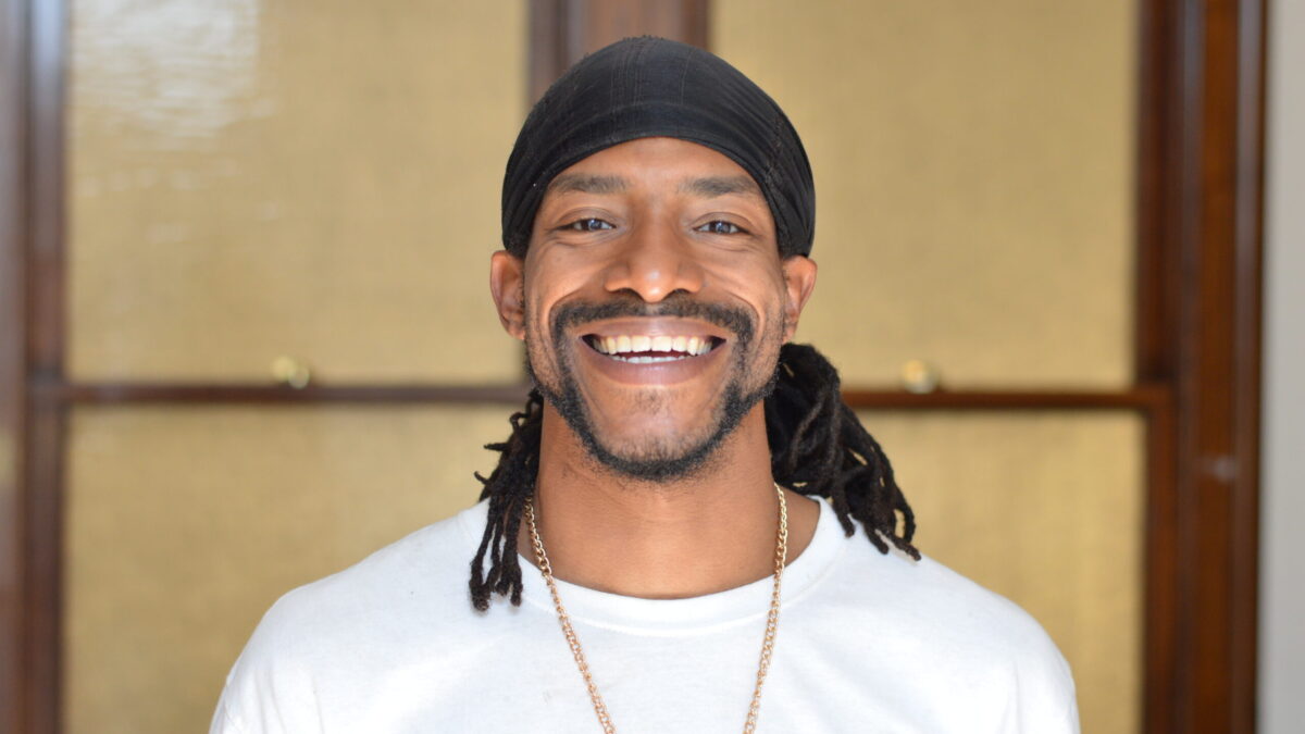 An adult with a short black beard and mustache and dark brown dreadlocks wearing a head scarf smiling in profile